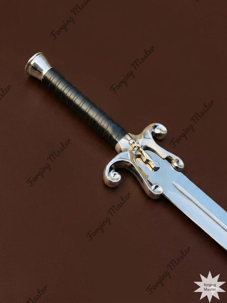 May include: A silver-toned sword featuring a black wrapped handle, a metallic pommel, and a crossguard detailed with a small skull emblem. The blade is polished steel with a central fuller, extending from a guard with curved, scroll-like quillons.