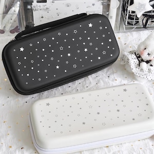 May include: Two rectangular cases for electronic devices. One is black with white stars and dots, the other is white with gray stars and dots. Both have zippers and a handle on the black case. The white case has a red and white charm.