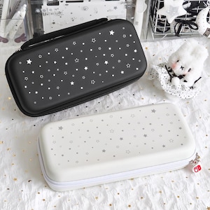 May include: Two rectangular cases for electronic devices. One is black with white stars and dots, the other is white with gray stars and dots. Both have zippers and a handle on the black case. The white case has a red and white charm.