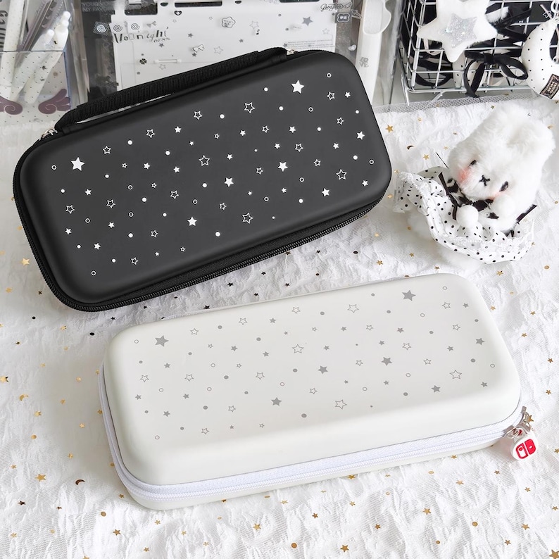 May include: Two rectangular cases for electronic devices. One is black with white stars and dots, the other is white with gray stars and dots. Both have zippers and a handle on the black case. The white case has a red and white charm.