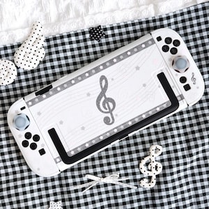May include: White Nintendo Switch console with a musical theme. The console features a gray treble clef design, star accents, and a gray border. The controllers have gray thumbsticks and black buttons. The console rests on a black and white checkered fabric.