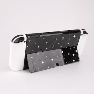 May include: Black and white Nintendo Switch console with a stand. The console is black with a white star pattern and white controllers. The stand is also black with a white star pattern. The console is designed to be used with the stand.
