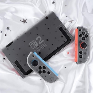 May include: A gray Nintendo Switch console and two controllers with a star pattern. The console has the text "Nintendo Switch 2". One controller is orange, the other is blue.