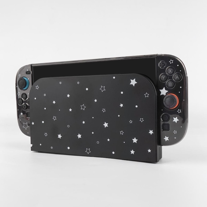 May include: A black gaming console with a matching stand. The stand is decorated with white stars and dots. The console has black controllers with blue and orange accents. The background is plain white.