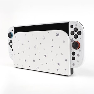 May include: A white Nintendo Switch dock and controller with a star pattern. The dock is white with a gray star pattern. The controller is white with black buttons and a red joystick. The Switch is black.