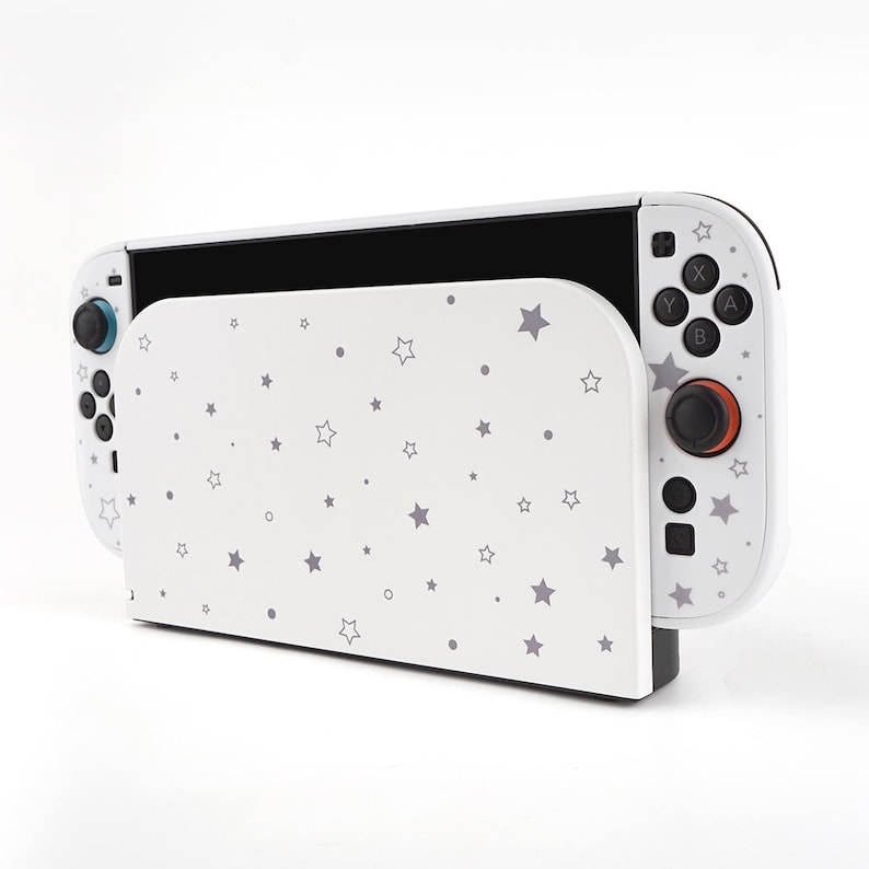 May include: A white Nintendo Switch dock and controller with a star pattern. The dock is white with a gray star pattern. The controller is white with black buttons and a red joystick. The Switch is black.