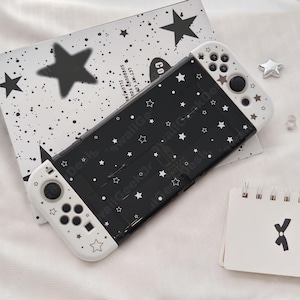 May include: Black and white Nintendo Switch case with a star pattern. The case has white controllers with star details. A matching black and white star-patterned card is behind the Switch. A small notepad with a black X is on the right.