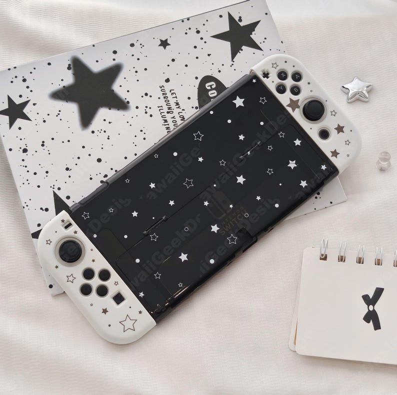 May include: Black and white Nintendo Switch case with a star pattern. The case has white controllers with star details. A matching black and white star-patterned card is behind the Switch. A small notepad with a black X is on the right.