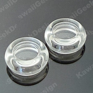 May include: Two clear, circular glass objects with a hole in the center. The objects are transparent, allowing light to pass through. They are sitting on a reflective surface, creating a mirrored effect. The design is simple and modern.