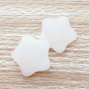 Op de afbeelding: Twee witte, stervormige objecten worden getoond tegen een lichtbruine achtergrond met houtnerf. De objecten hebben een gladde, matte afwerking en afgeronde randen. De sterren zijn qua grootte en vorm vergelijkbaar.