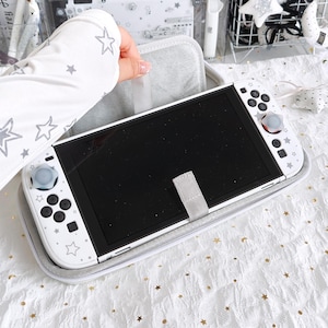 May include: White Nintendo Switch console with a gray case. The console has a black screen and white controllers decorated with star patterns. The case is gray with a gray strap to secure the console. The controllers have gray and red accents.