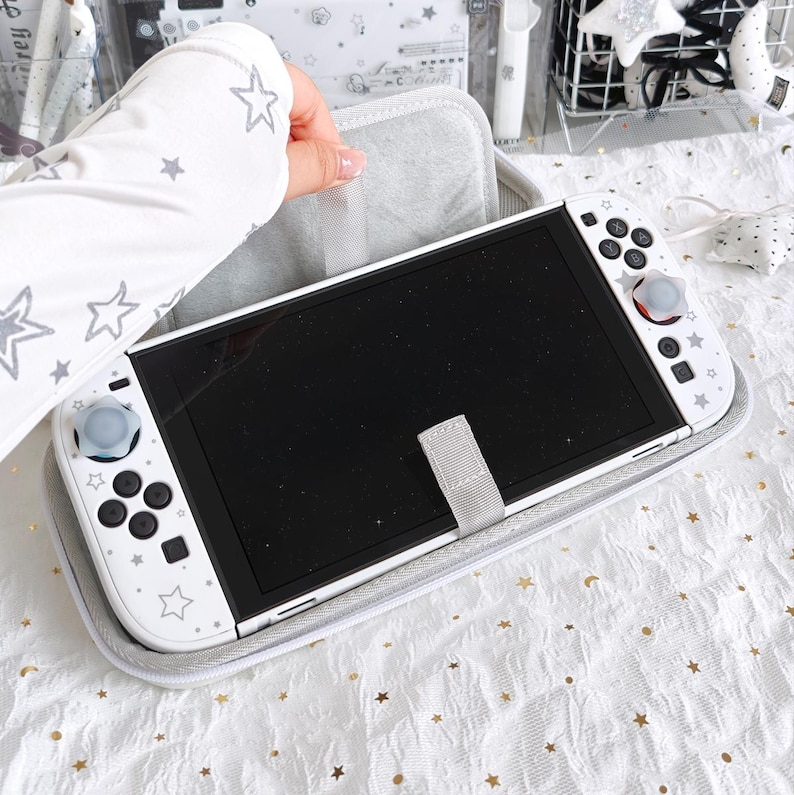 May include: White Nintendo Switch console with a gray case. The console has a black screen and white controllers decorated with star patterns. The case is gray with a gray strap to secure the console. The controllers have gray and red accents.