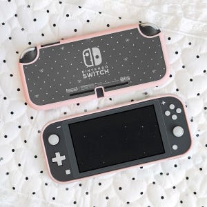 Könnte beinhalten: Eine tragbare Nintendo Switch Lite Spielkonsole mit dunkelgrauem Gehäuse und hellrosa Rahmen. Die Rückseite zeigt ein Muster aus weißen Punkten und kleinen Schleifen, während die Vorderseite einen schwarzen Bildschirm und graue Tasten aufweist.