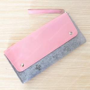 May include: A pink and gray clutch with a wrist strap. The pink flap is secured with two silver snaps and covers a gray felt body with a controller design. The clutch has a cross, circle, and button design.