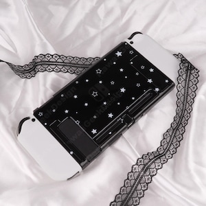May include: A black and white Nintendo Switch case with a star pattern. The case has white accents and a built-in stand. A black lace ribbon is draped across the case. The text "Kawaii Geek Design" is printed on the case.