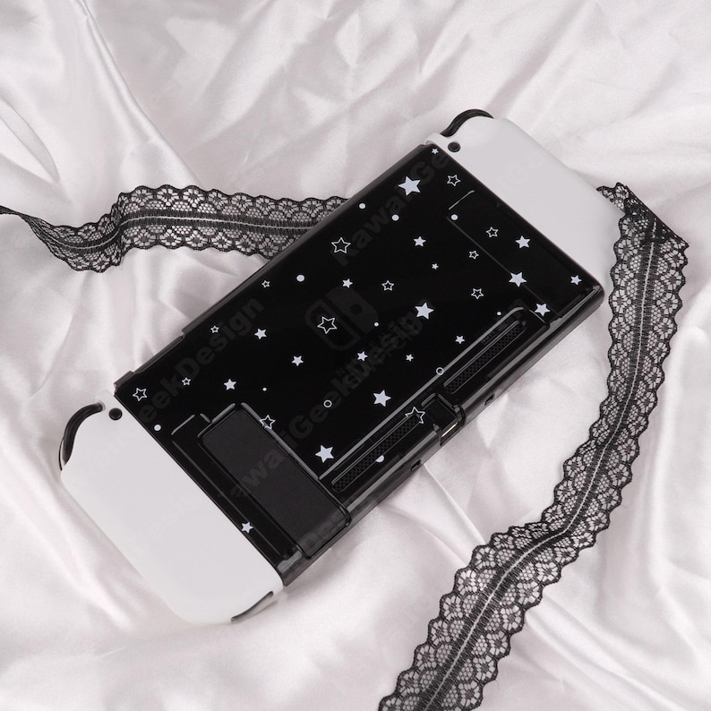 May include: A black and white Nintendo Switch case with a star pattern. The case has white accents and a built-in stand. A black lace ribbon is draped across the case. The text "Kawaii Geek Design" is printed on the case.
