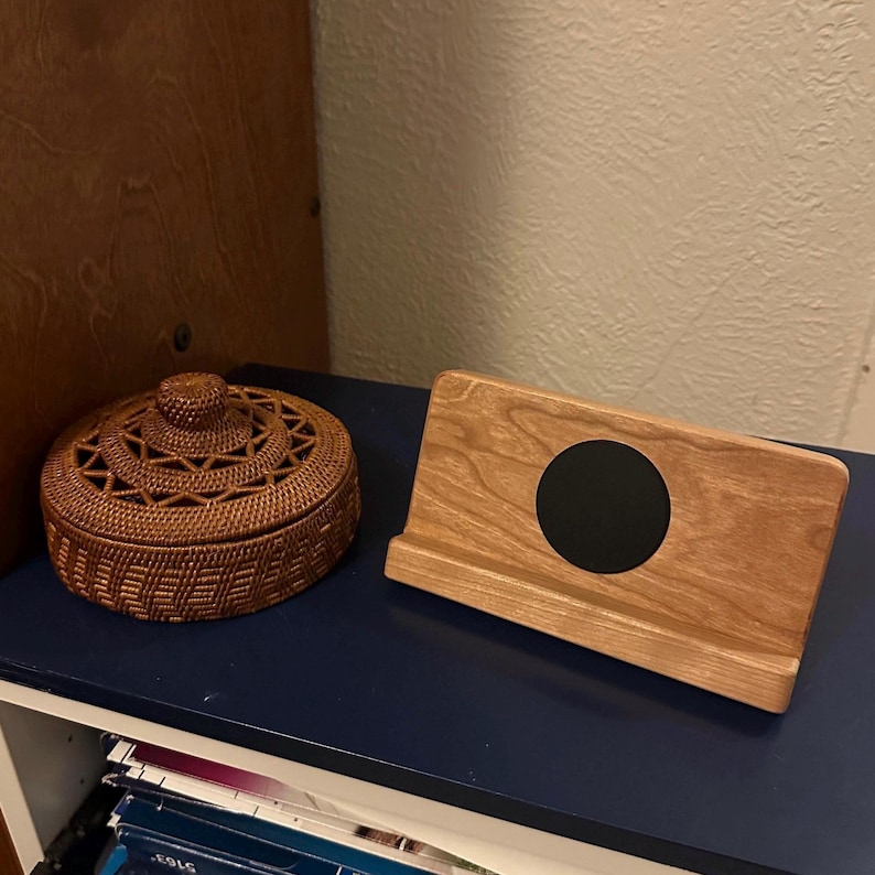 Puede incluir: Una cesta marr&oacute;n tejida con tapa y un soporte de tel&eacute;fono de madera con un c&iacute;rculo negro sobre una estanter&iacute;a azul. La cesta tiene un dise&ntilde;o circular texturizado. El soporte del tel&eacute;fono es de madera clara.
