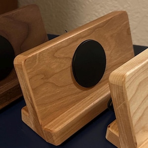Puede incluir: Soporte de tel&eacute;fono de madera con acabado natural y un acento circular negro. El soporte est&aacute; inclinado para una visualizaci&oacute;n &oacute;ptima y tiene una ranura para sostener un tel&eacute;fono o tableta. La veta de la madera es visible.