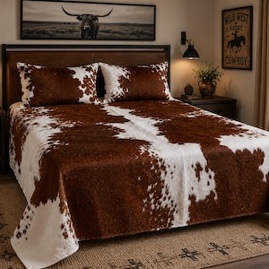 May include: A bedspread and two matching pillow shams featuring a brown and white cowhide print pattern. The bedding set covers a bed frame with a brown headboard, displaying a consistent spotted animal hide aesthetic across the fabric surface.