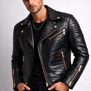 May include: Black leather jacket with a crocodile skin texture, featuring gold zippers and hardware. The jacket has a classic biker style with a notched collar and quilted shoulder details. The model is wearing a black shirt and black jeans.