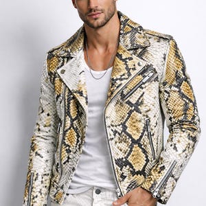 May include: A snakeskin-patterned leather jacket in shades of white, tan, and black. The jacket features a classic biker style with a notched collar, zippered pockets, and a front zipper closure. The model is wearing a white t-shirt and white jeans.