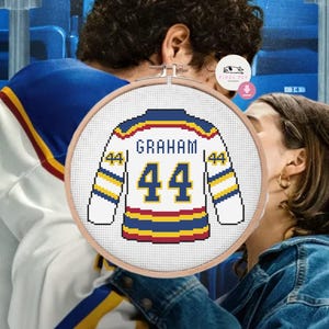 Könnte beinhalten: Ein Hockey-Trikot-Design im Kreuzstich in einem Holzreifen. Das Trikot ist weiß mit blauen, gelben und roten Akzenten und trägt den Namen "GRAHAM" und die Nummer "44". Das Design befindet sich vor einem verschwommenen Hintergrund von Personen und Stadionbestuhlung.