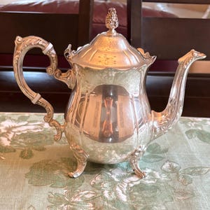 Peut inclure: Une théière argentée avec des détails ornés. La théière présente une anse incurvée, un bec verseur et un couvercle avec un fleuron décoratif. Le corps de la théière reflète l'environnement. Elle repose sur une nappe à motifs floraux.