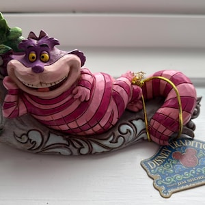 Può includere: Una statuina del Gatto del Cheshire di Disney Traditions. Il gatto, a strisce rosa e viola, è sdraiato con un ampio sorriso. Poggia su una base decorativa con un anello dorato per appenderlo. Oggetto da collezione.