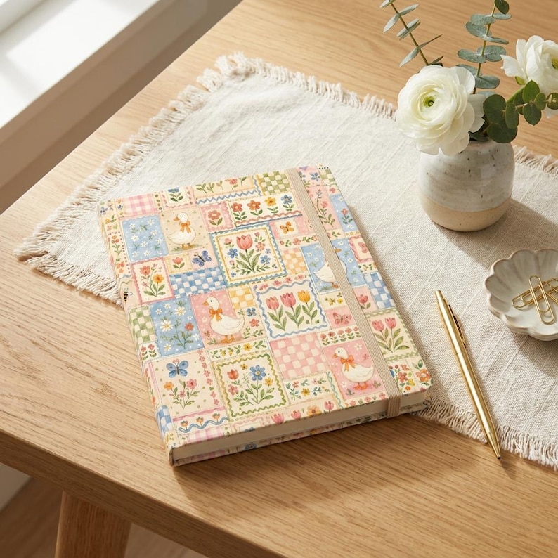 Puede incluir: Un diario de tapa dura con un dise&ntilde;o de patchwork con patos, flores y mariposas en colores pastel. El diario tiene un cierre el&aacute;stico y est&aacute; sobre una mesa de madera con un bol&iacute;grafo, flores y un plato decorativo.
