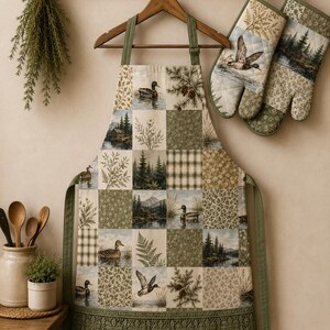 May include: A patchwork apron and matching oven mitts featuring a nature-themed print with ducks, pine trees, mountains, and floral patterns in shades of green, brown, and cream. The apron has olive green straps and a decorative lace trim at the hem.