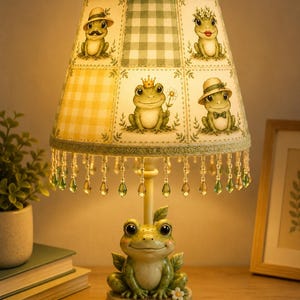 May include: A table lamp featuring a base shaped like a green frog with large eyes. The lampshade is decorated with a grid of illustrations of frogs wearing hats and crowns, accented by a green gingham pattern and hanging green crystal beads.