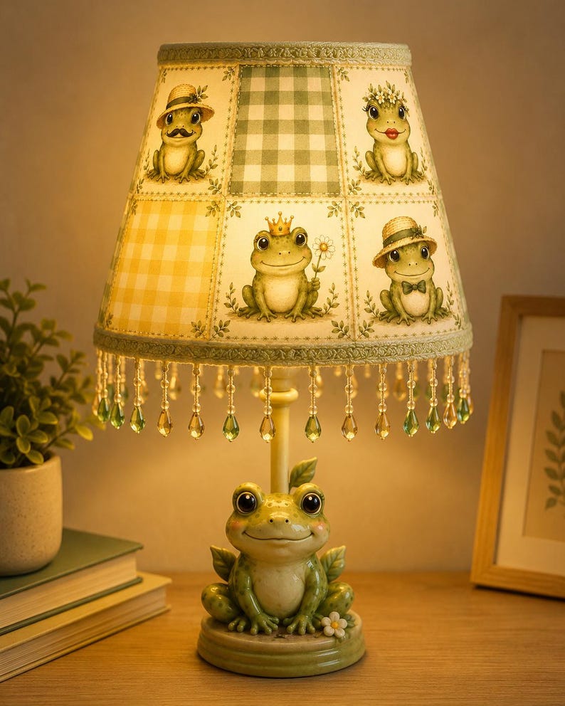 May include: A table lamp featuring a base shaped like a green frog with large eyes. The lampshade is decorated with a grid of illustrations of frogs wearing hats and crowns, accented by a green gingham pattern and hanging green crystal beads.