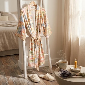 Puede incluir: Una bata con estampado floral cuelga de una escalera blanca, con zapatillas a juego debajo. La bata presenta un dise&ntilde;o de patchwork con colores pastel y motivos florales y de conejos. Una taza de t&eacute;, una vela y ramitas de lavanda est&aacute;n sobre una mesa auxiliar.