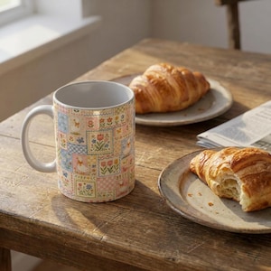 Puede incluir: Una taza de cer&aacute;mica con un dise&ntilde;o de patchwork con motivos florales y animales en colores pastel. La taza est&aacute; sobre una mesa de madera con cruasanes en platos.