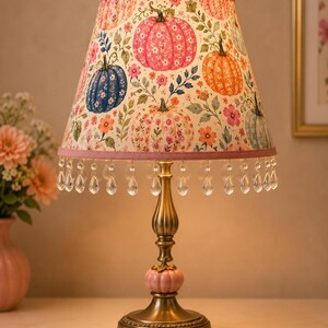 May include: A table lamp with a brass-toned metal base featuring a small pink pumpkin accent. The conical lampshade displays a pattern of colorful pumpkins and flowers, finished with a pink fabric trim and hanging clear crystal-like teardrop beads.