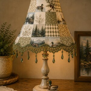 May include: A table lamp with a patchwork fabric shade featuring nature scenes of ducks, pine trees, and cabins. The shade has a scalloped green lace trim with hanging green teardrop beads. The base is a distressed cream colour with a mallard duck figure.