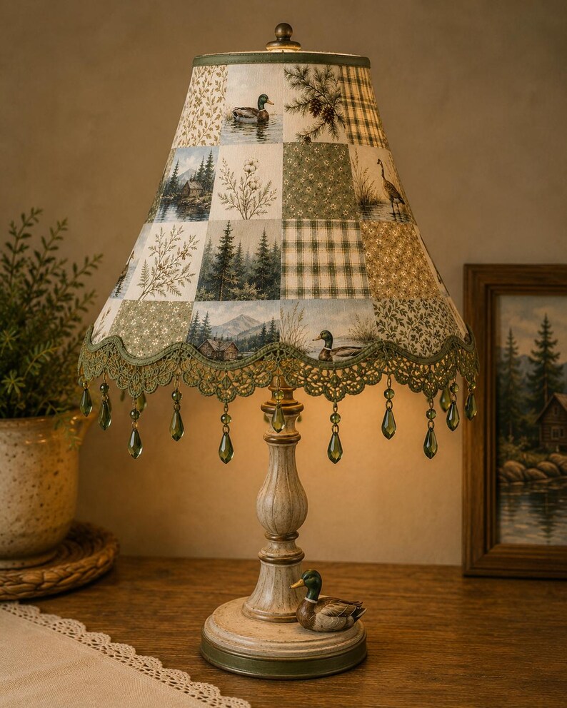 May include: A table lamp with a patchwork fabric shade featuring nature scenes of ducks, pine trees, and cabins. The shade has a scalloped green lace trim with hanging green teardrop beads. The base is a distressed cream colour with a mallard duck figure.