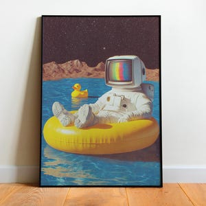 May include: A framed art print depicts an astronaut with a television for a head, relaxing on a yellow inflatable ring in a pool of water. A rubber duck floats nearby. The background shows a starry sky and a mountain range.