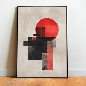 May include: Abstract art print in a black frame. The artwork features a large red circle and geometric shapes in black and red against a neutral background. The framed art is leaning against a white wall on a wooden floor.