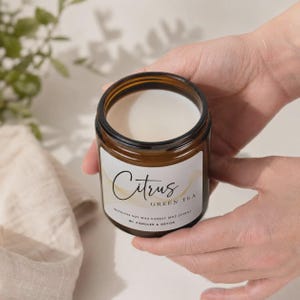 May include: A wickless soy wax candle in an amber glass jar with a white label featuring the text Citrus Green Tea and 8oz (226g). The candle has a smooth white wax surface and is contained within a cylindrical glass vessel with a black rim.