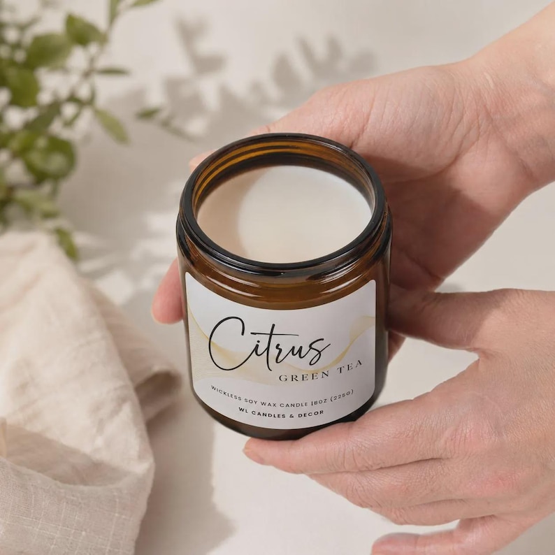May include: A wickless soy wax candle in an amber glass jar with a white label featuring the text Citrus Green Tea and 8oz (226g). The candle has a smooth white wax surface and is contained within a cylindrical glass vessel with a black rim.