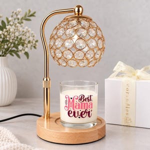 May include: A gold-toned candle warmer lamp with a spherical shade covered in clear crystals, mounted on a light wood base. A glass candle jar sits on the base, featuring the pink and dark red text "Best Mama ever" printed on the front.