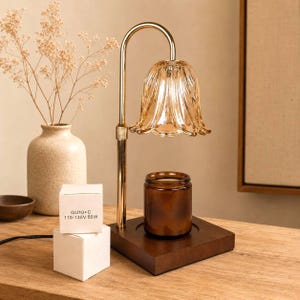 May include: A gold-toned metal candle warmer lamp with a curved arm and a fluted, amber-tinted glass shade. The lamp features a square dark wood base holding a brown glass candle jar. A white box displays the text GU10+C 110-130V 50W.