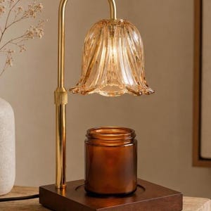May include: A candle warmer lamp featuring a gold-toned metal stand with a curved neck and a fluted, amber-colored glass shade. The lamp sits on a square, dark wood base, positioned directly above a cylindrical amber glass candle jar.