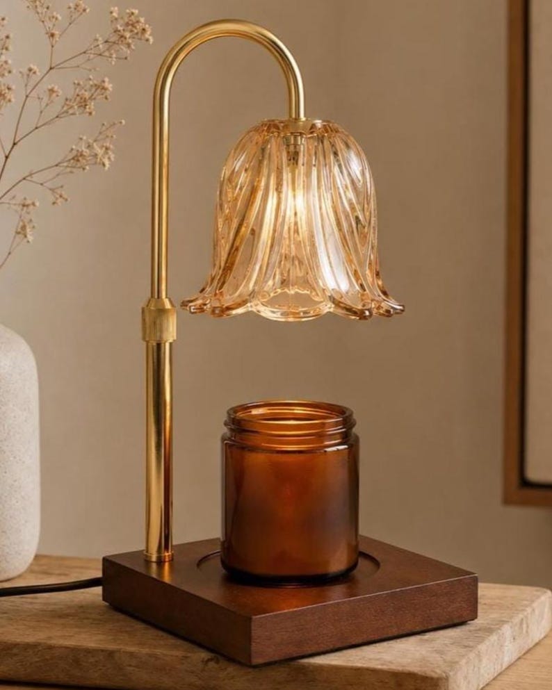 May include: A candle warmer lamp featuring a gold-toned metal stand with a curved neck and a fluted, amber-colored glass shade. The lamp sits on a square, dark wood base, positioned directly above a cylindrical amber glass candle jar.