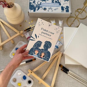 Puede incluir: Un kit de pintura creativo con una guía titulada "Paint your partner". El kit incluye pequeños caballetes de madera, tubos de pintura, paletas de pintura con varios colores, pinceles y lienzos en blanco. También se ve una caja con texto en francés.