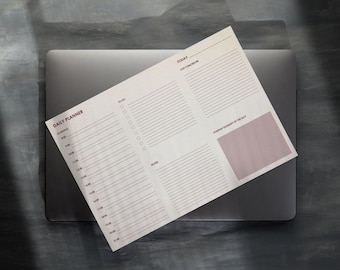 Könnte beinhalten: Ein Tagesplaner mit hellrosa und weißem Design, der auf einem grauen Laptop liegt. Der Planer enthält Abschnitte für To-Do-Listen, Terminplanung und einen Bereich für einen positiven Gedanken des Tages. Die Worte "Daily Planner" sind oben aufgedruckt.