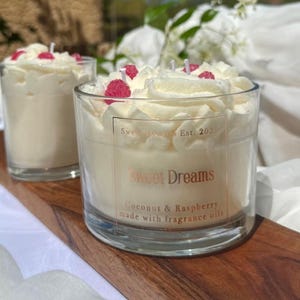 May include: Two clear glass candles topped with white wax piped to resemble whipped cream and decorated with small pink raspberry-shaped wax pieces. The front label reads Sweet Dreams Est. 2025, Coconut & Raspberry made with fragrance oils.