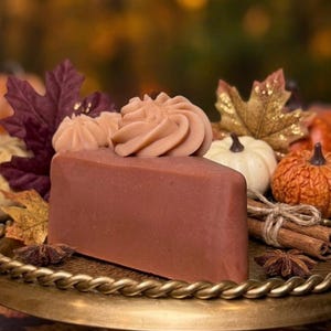 May include: A triangular slice of brown soap shaped like a cake, topped with two piped swirls of light brown soap frosting. The soap is displayed on a gold-toned metal pedestal tray alongside decorative autumn leaves, small pumpkins, and cinnamon sticks.
