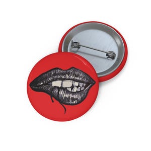 以下が含まれることがあります： 2つの丸いバッジ。1つは赤い背景に口の白黒イラストが描かれています。もう1つは赤色で、裏側に銀色のピン留めが付いています。